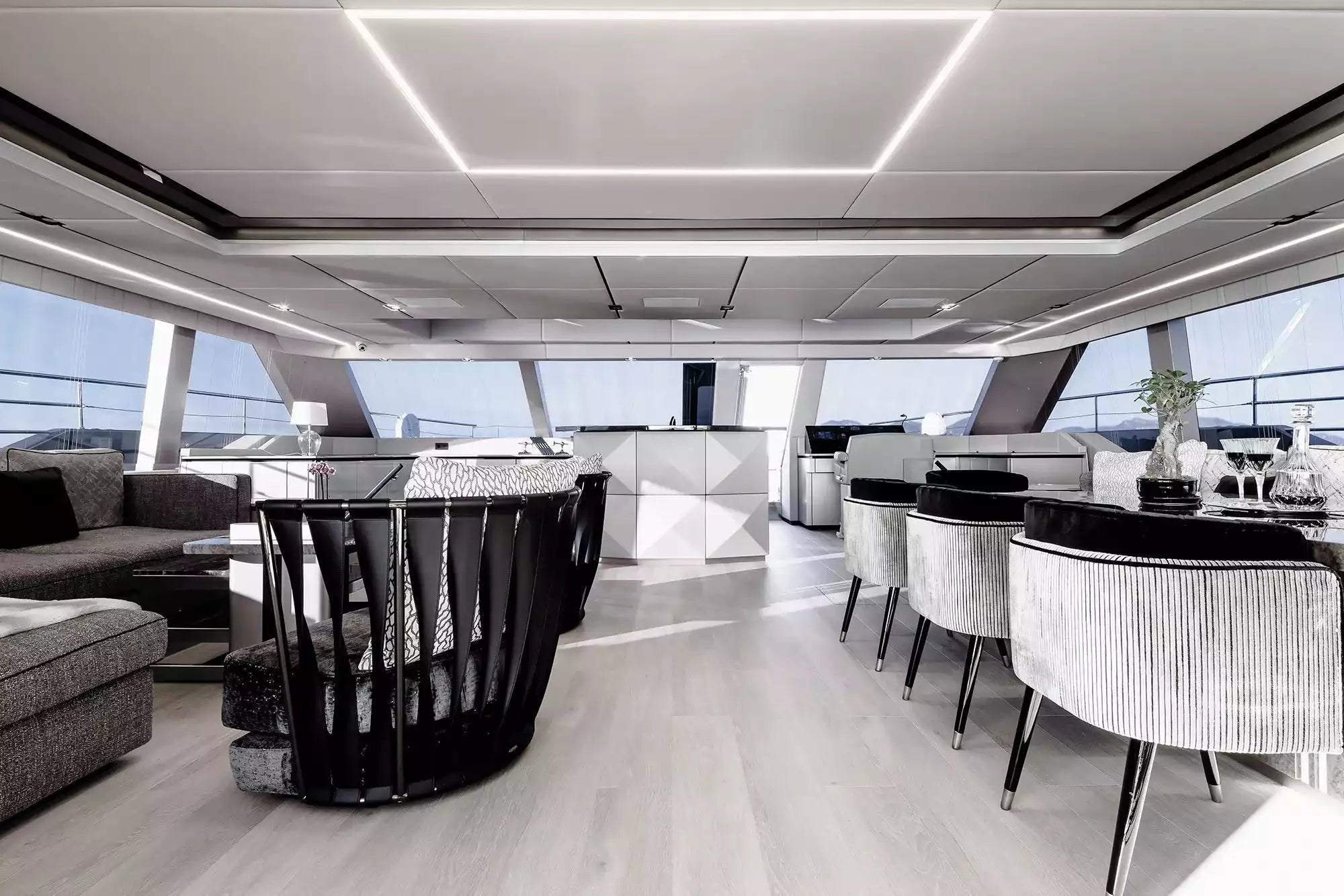 A luxury yacht interior featuring a modern living and dining area with light gray wood flooring, white and gray furniture, and large windows looking out to the sea.