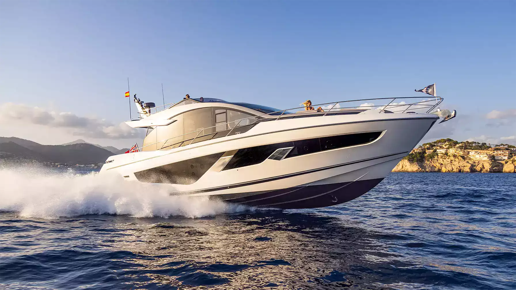 A luxury motor yacht speeds across deep blue water, creating a white spray wake. The yacht is white with dark gray and black accents, featuring a modern open-top design.