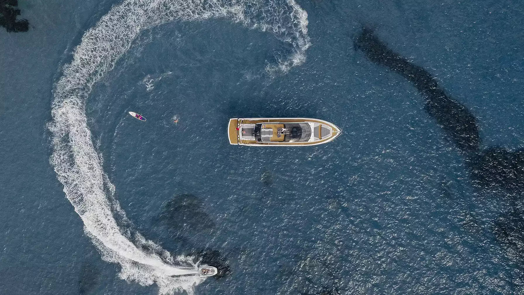 Aerial view of a luxury motor yacht on deep blue water, with a wake trail created by a jet ski nearby and a small boat at the bottom edge.