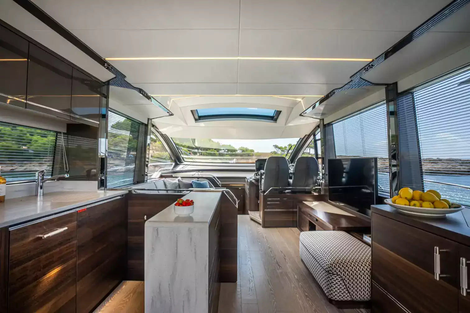 The interior of a luxury yacht or motor yacht cabin featuring a modern galley kitchen with dark wood cabinets, a marble-topped island, a seating area with a patterned ottoman, and large windows looking out onto water.