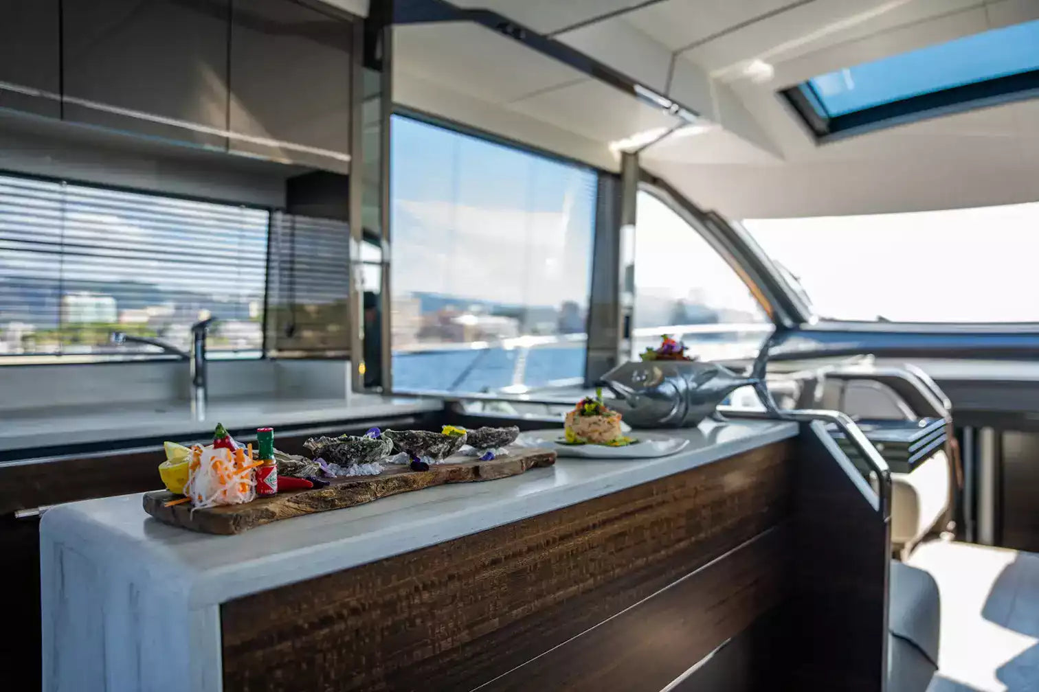 A modern yacht galley interior with a white countertop, dark wood cabinetry, large windows overlooking water, and plated food arranged on the counter.
