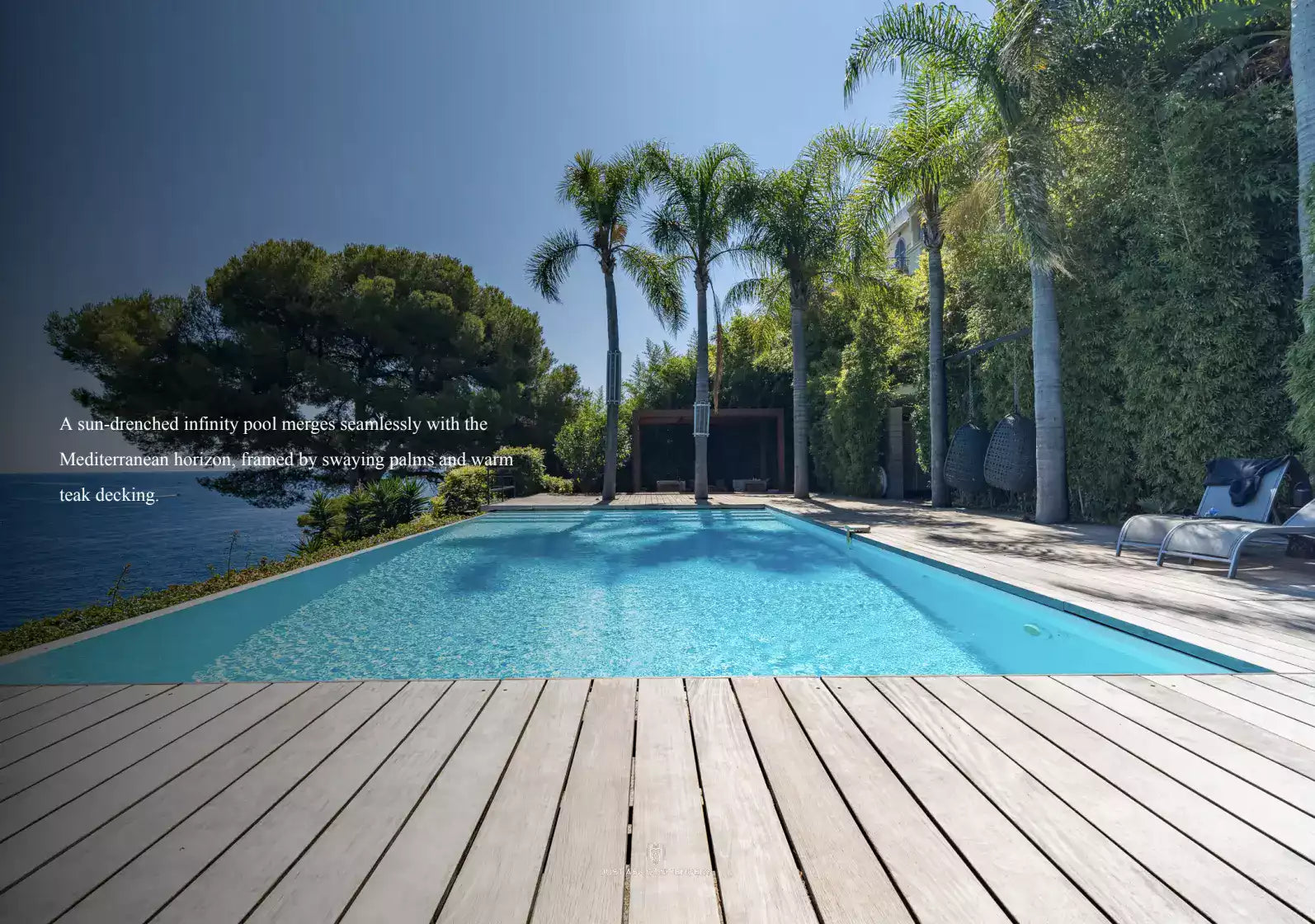 Infinity pool with wooden deck, palm trees, and Mediterranean-style house.