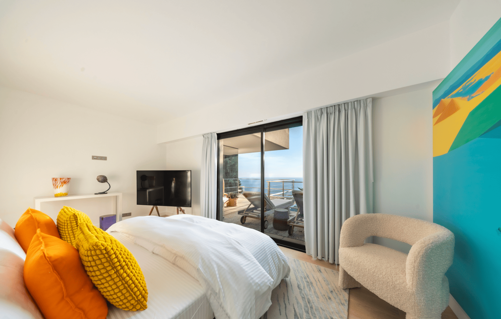 Modern bedroom with a bed, colorful pillows, and a view of the ocean.