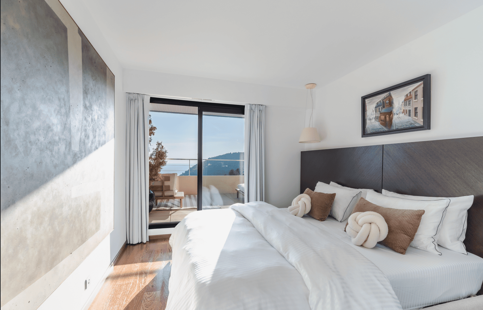 Modern bedroom with a large bed, pillows, and a view of the ocean through a window.