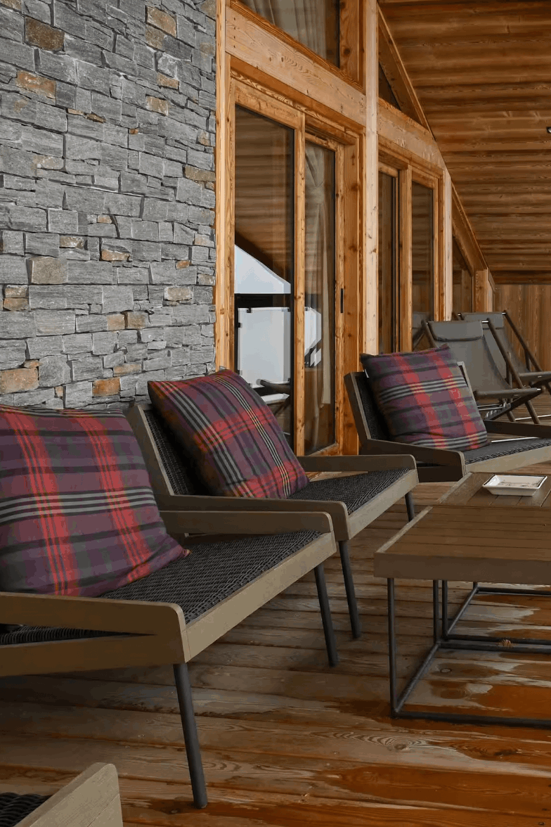 A wooden deck balcony with outdoor seating, including two gray armchairs with red and black plaid throw pillows, a matching wooden coffee table, and a rustic wood log side table. The background features a gray stone wall, wooden posts, and a snowy mountain view visible through the glass windows.