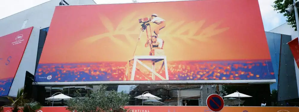 A large outdoor digital billboard mounted on a building, displaying a warm-toned graphic with a person standing on a ladder against a red, orange, and yellow background. The billboard is situated in an urban setting with trees, umbrellas, and surrounding structures visible.