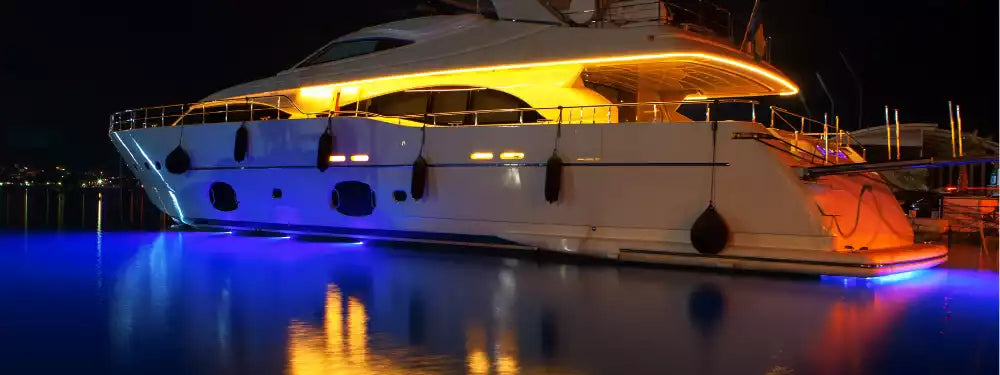 A luxury yacht docked at night in a marina, illuminated with warm yellow lights on the upper deck and cool blue LED lights along the hull, reflecting on the water.