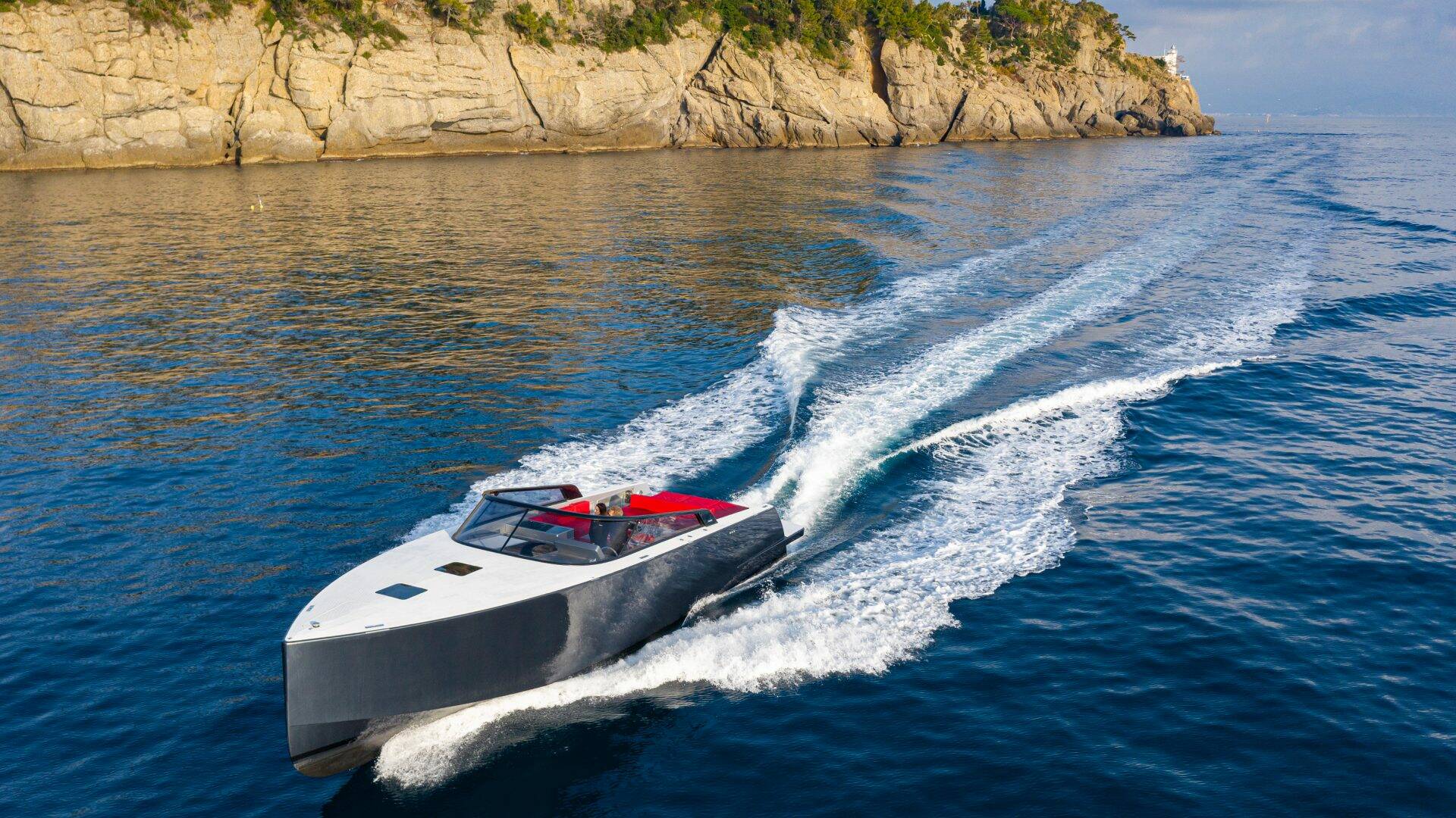 White speedboat with red interior cutting through blue water near a rocky coastline.